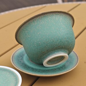 K&ouml;nnte beinhalten: Eine blaugr&uuml;ne Keramik-Teetasse, die schr&auml;g auf einer passenden Untertasse steht. Die Tasse hat eine gesprenkelte, matte Oberfl&auml;che mit einem dunklen Rand und einem wei&szlig;en runden Boden. Die Untertasse zeigt eine blaugr&uuml;ne Glasur mit dunklem Rand.