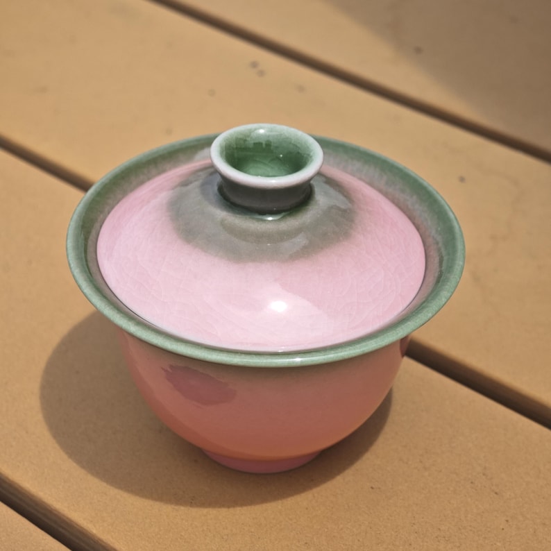 Puede incluir: Un cuenco de t&eacute; gaiwan de cer&aacute;mica con tapa a juego, que presenta un color base rosa suave que transiciona hacia un esmalte verde salvia apagado alrededor del borde y el pomo central de la tapa, con un acabado liso, brillante y forma redondeada.