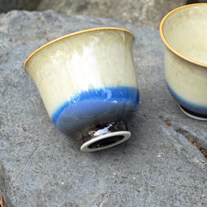 May include: Two ceramic tea cups with a gradient design. The cups have a cream-colored top, transitioning to blue, and a dark brown base. The rims are outlined in gold. The cups are on a gray stone surface.