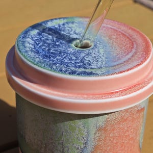 Pu&ograve; includere: Un bicchiere in ceramica con coperchio. Il coperchio &egrave; diviso in sezioni blu e rosse, con un foro per una cannuccia. Il corpo del bicchiere ha un aspetto strutturato e screziato con sfumature verdi e arancioni. Una cannuccia trasparente &egrave; inserita nel coperchio.