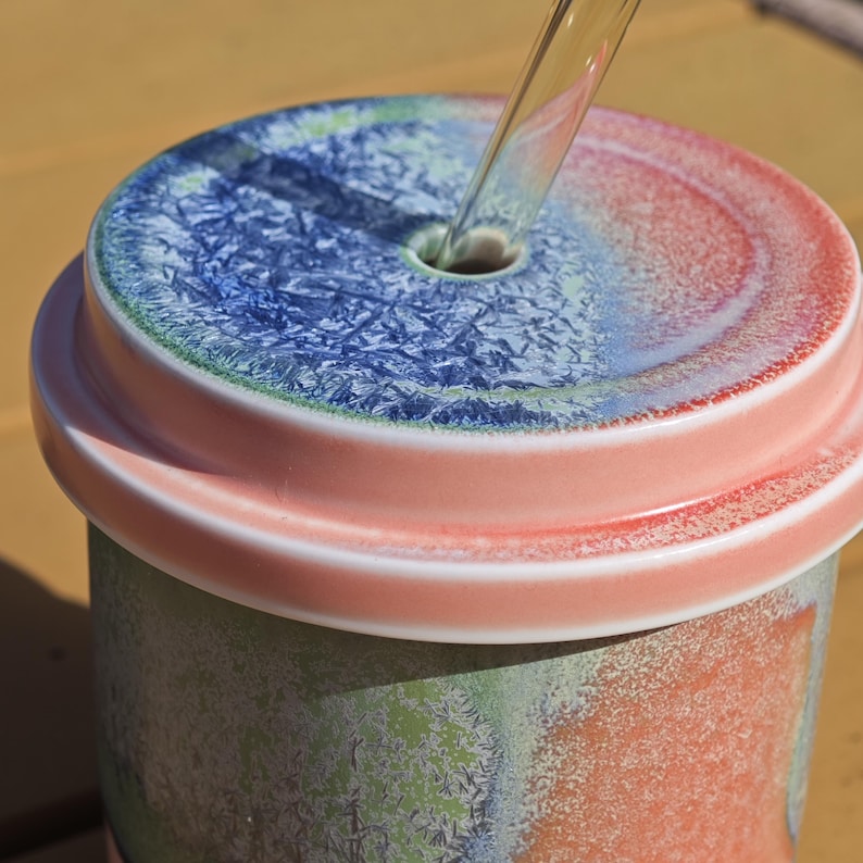 Pu&ograve; includere: Un bicchiere in ceramica con coperchio. Il coperchio &egrave; diviso in sezioni blu e rosse, con un foro per una cannuccia. Il corpo del bicchiere ha un aspetto strutturato e screziato con sfumature verdi e arancioni. Una cannuccia trasparente &egrave; inserita nel coperchio.