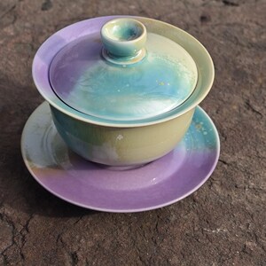 May include: A ceramic gaiwan tea set with a lid, bowl, and saucer. The set features a gradient of colors, including purple, blue, and green. The gaiwan is designed for brewing and serving tea.