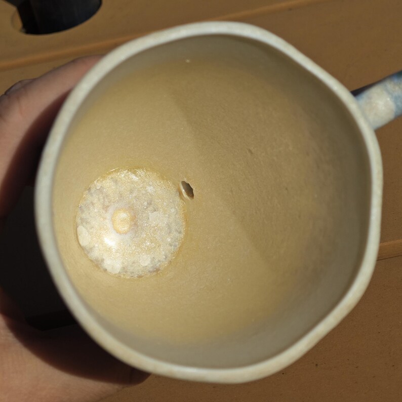 May include: A handmade ceramic mug with a light beige interior and a white rim. The mug has a small hole on the side and a textured bottom. The handle is partially visible, with blue and white stripes. The mug is empty.