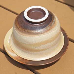 May include: A ceramic bowl turned upside down on a matching saucer. The bowl features a gradient of dark brown at the top, transitioning into a creamy beige with horizontal textured bands. The base rim is white, and the saucer is dark brown.
