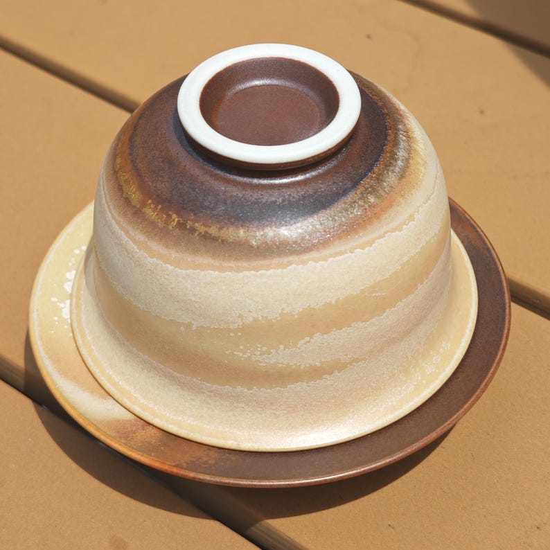 May include: A ceramic bowl turned upside down on a matching saucer. The bowl features a gradient of dark brown at the top, transitioning into a creamy beige with horizontal textured bands. The base rim is white, and the saucer is dark brown.