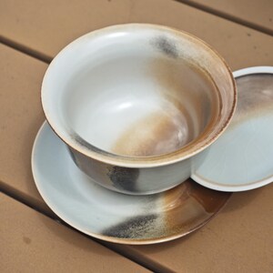 May include: A ceramic tea bowl with a matching saucer and lid, featuring a light blue-grey base glaze with blended brown and charcoal black accents. The bowl has a wide, open rim and a smooth, rounded interior, while the saucer has a flat base.