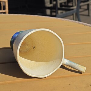 May include: A handmade ceramic mug with a wide, open mouth and a small handle. The interior is a warm, light yellow, while the exterior is a soft, muted blue and white. The mug is resting on a wooden surface.