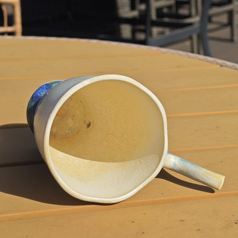May include: A handmade ceramic mug with a wide, open mouth and a small handle. The interior is a warm, light yellow, while the exterior is a soft, muted blue and white. The mug is resting on a wooden surface.