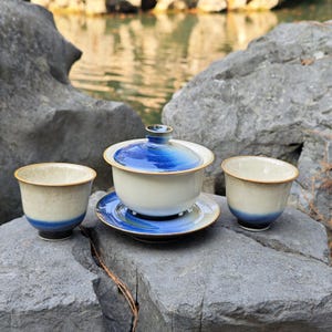 May include: A ceramic tea set consisting of a lidded gaiwan on a saucer and two matching tea cups. The pieces feature a gradient glaze transitioning from a creamy off-white base to a deep, vibrant blue at the rim and lid, with thin brown edges.