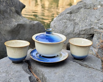 May include: A ceramic tea set consisting of a lidded gaiwan on a saucer and two matching tea cups. The pieces feature a gradient glaze transitioning from a creamy off-white base to a deep, vibrant blue at the rim and lid, with thin brown edges.