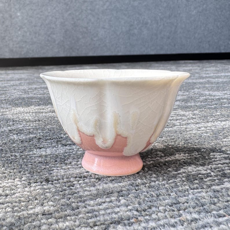 Pode incluir: Uma pequena tigela de cer&acirc;mica com esmalte branco rachado e base rosa. A tigela tem uma borda alargada e uma base de pedestal. O esmalte branco escorre para uma faixa rosa, criando um efeito visual &uacute;nico. A tigela &eacute; provavelmente feita &agrave; m&atilde;o.