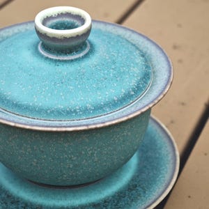 K&ouml;nnte beinhalten: Ein Gaiwan-Teeservice aus Keramik, bestehend aus einer Schale, einem Deckel mit zentralem Knauf und einer passenden Untertasse. Das Set ist mit einer gesprenkelten blaugr&uuml;nen und t&uuml;rkisfarbenen Glasur und dunkelbraunen R&auml;ndern versehen.