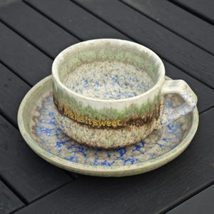 May include: A ceramic teacup and saucer set with a speckled design in shades of green, brown, and blue. The cup has a handle and the words "Make it sweet" are written on the side in gold lettering. The saucer has a matching pattern.