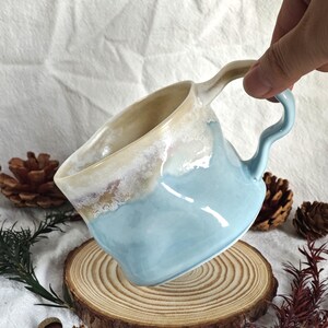 May include: A handmade ceramic mug with a unique, wavy handle. The mug features a light blue exterior with a creamy, textured rim. The mug is displayed on a wooden slice, showcasing its artisanal quality. Perfect for coffee or tea.