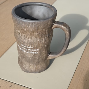 Puede incluir: Una taza alta de cer&aacute;mica con un exterior marr&oacute;n texturizado similar a la corteza y un interior gris liso. La taza cuenta con un asa curva y texto impreso en blanco que dice: Michael - Morning Person (After Coffee). Acabado moteado.