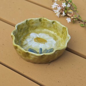 May include: A small, round, ceramic bowl with a scalloped edge. The bowl is painted with a large white daisy with a yellow center and blue accents on a light green background. The bowl is on a brown wooden surface.