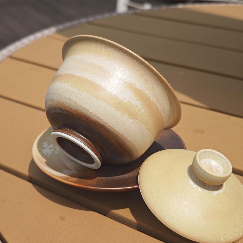 May include: A ceramic tea bowl with a lid and saucer, featuring a gradient glaze that transitions from a creamy beige at the rim to a deep, dark brown at the base. The bowl is tilted on its saucer, revealing a white interior rim and a smooth finish.