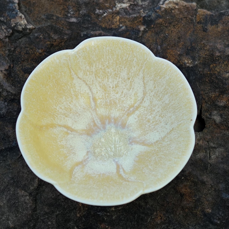 May include: A small, flower-shaped ceramic dish with a creamy yellow glaze. The dish has a slightly textured surface and a delicate, petal-like edge. The interior features subtle, radiating patterns.
