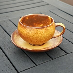 May include: A ceramic teacup and saucer set. The cup and saucer are a warm, speckled brown and yellow. The cup has a rounded shape with a handle. The saucer is round and flat. The set is on a dark gray wooden surface.