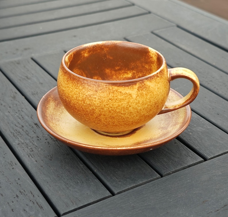 May include: A ceramic teacup and saucer set. The cup and saucer are a warm, speckled brown and yellow. The cup has a rounded shape with a handle. The saucer is round and flat. The set is on a dark gray wooden surface.