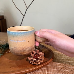 May include: A ceramic mug with a brown and blue gradient design, filled with a dark liquid. The mug has the text "Feeling Peachy" and is held by a person. A wooden handle and a pine cone are also visible.