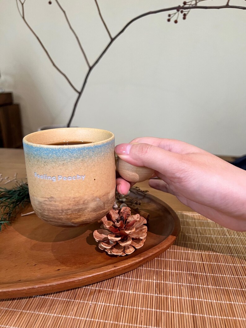 May include: A ceramic mug with a brown and blue gradient design, filled with a dark liquid. The mug has the text "Feeling Peachy" and is held by a person. A wooden handle and a pine cone are also visible.