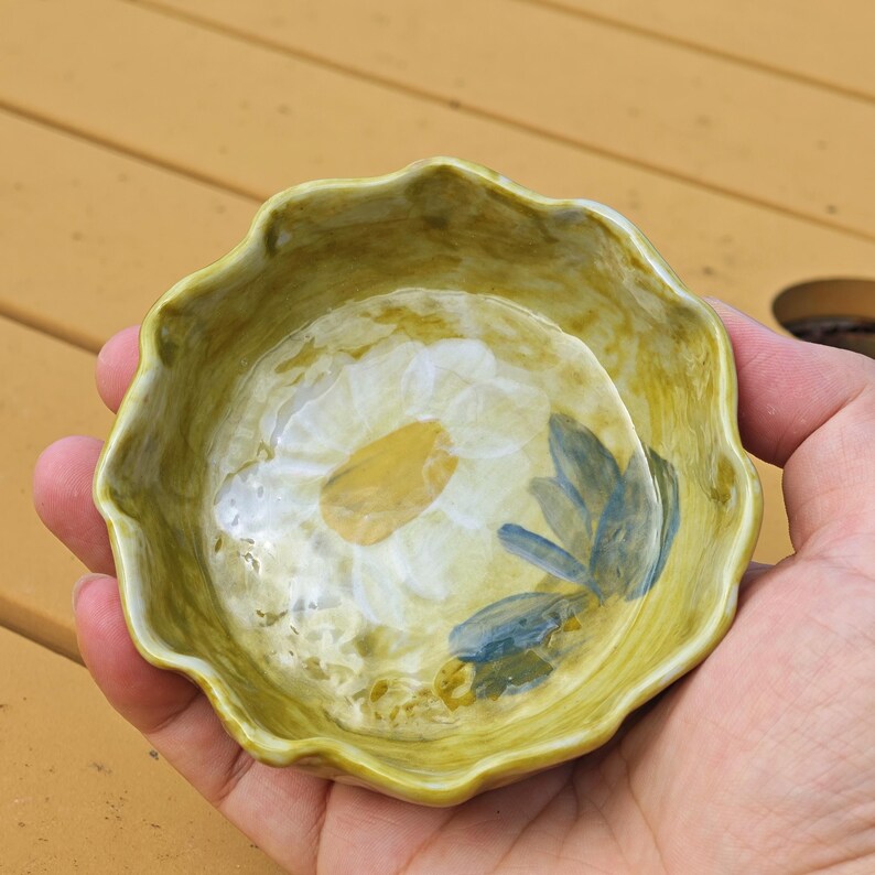 May include: A small, hand-painted ceramic bowl with a scalloped edge. The bowl features a white daisy design with yellow and blue accents. The bowl is held in a person's hand.