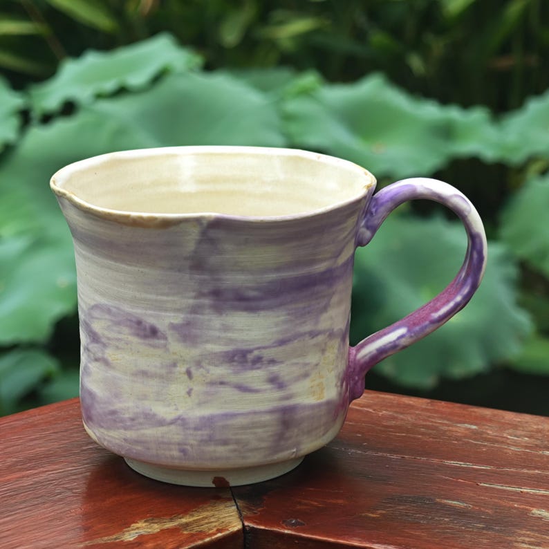 May include: A handmade ceramic mug with a unique marbled design in shades of purple and cream. The mug features a rounded body, a sturdy handle, and a slightly flared rim. The mug is perfect for enjoying coffee, tea, or any favorite beverage.