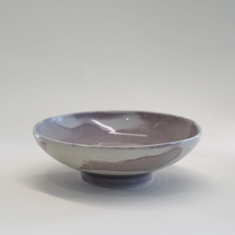 May include: A ceramic bowl with a wide, shallow design. The bowl features a muted color palette with shades of lavender, gray, and white. It has a small, circular base and a smooth, glossy finish. The bowl is suitable for serving food.