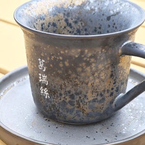 May include: A dark blue ceramic mug with a handle, featuring a speckled gold and black design. The mug sits on a matching saucer. White Japanese characters are printed on the side of the mug.