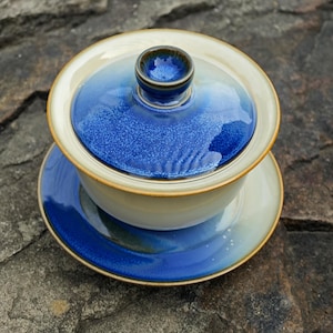 May include: A ceramic gaiwan tea set consisting of a bowl, a lid with a central knob, and a saucer. The set features a gradient glaze transitioning from a deep, vibrant blue at the top and edges to a soft, creamy beige at the base of the bowl.