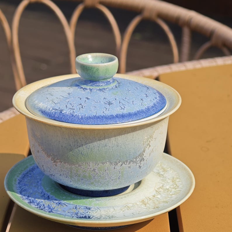 May include: A ceramic tea set with a blue and green color scheme. The set includes a lidded bowl, a small lid handle, and a saucer. The bowl and saucer have a textured surface with a light blue and green color gradient. The lid is light blue with a darker blue rim.