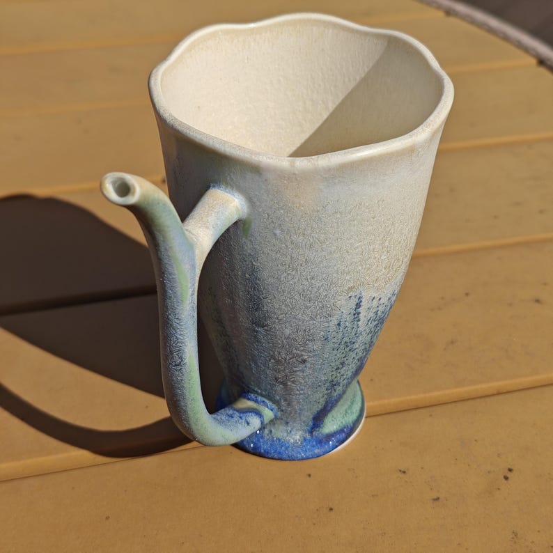 May include: A handmade ceramic mug with a unique spout and handle. The mug features a wavy rim and a color gradient, transitioning from off-white at the top to blue at the base. The handle and spout are a light green color.