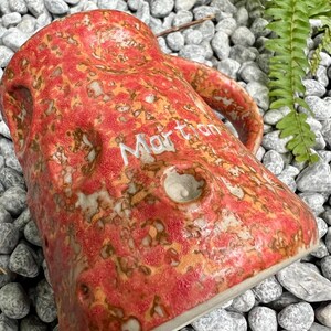 Op de afbeelding: Een handgemaakte keramische mok met een getextureerde, rood-oranje glazuur en het woord "Martian" in het wit. De mok heeft een unieke, organische vorm met een handvat en een klein gaatje. De mok staat tegen een achtergrond van grijze stenen en groen gebladerte.