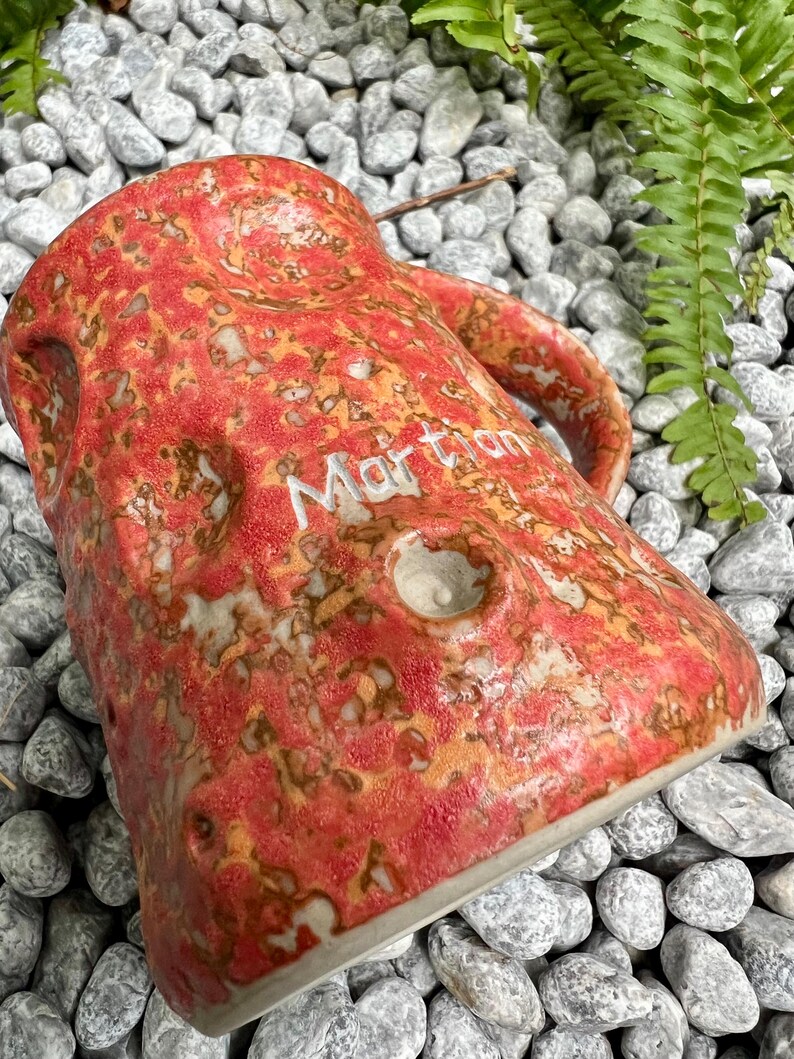 Op de afbeelding: Een handgemaakte keramische mok met een getextureerde, rood-oranje glazuur en het woord "Martian" in het wit. De mok heeft een unieke, organische vorm met een handvat en een klein gaatje. De mok staat tegen een achtergrond van grijze stenen en groen gebladerte.