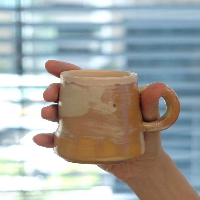May include: A handmade ceramic mug with a rounded handle, held in a person's hand. The mug features a warm color palette of tan and brown hues, with a slightly textured surface. The mug's design is simple and rustic, suggesting a focus on craftsmanship.