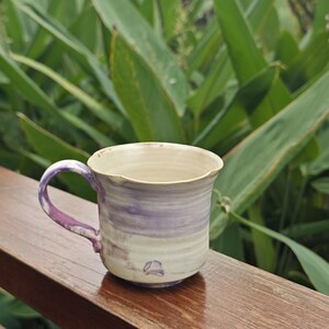 May include: A handmade ceramic mug with a purple and white swirl design. The handle and rim are also purple. The mug is sitting on a wooden surface with green foliage in the background. The mug has a unique, organic shape.