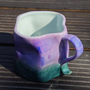 May include: A handmade ceramic mug with a unique, irregular shape. The mug features a gradient of colors, transitioning from green at the base to purple and lavender at the top, with a white interior. The handle is also purple.