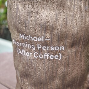 Puede incluir: Una taza cil&iacute;ndrica de color marr&oacute;n con una textura rugosa similar a la corteza de un &aacute;rbol. La superficie presenta un texto grabado en blanco que dice: Michael - Morning Person (After Coffee). El dise&ntilde;o imita un tronco con surcos verticales.