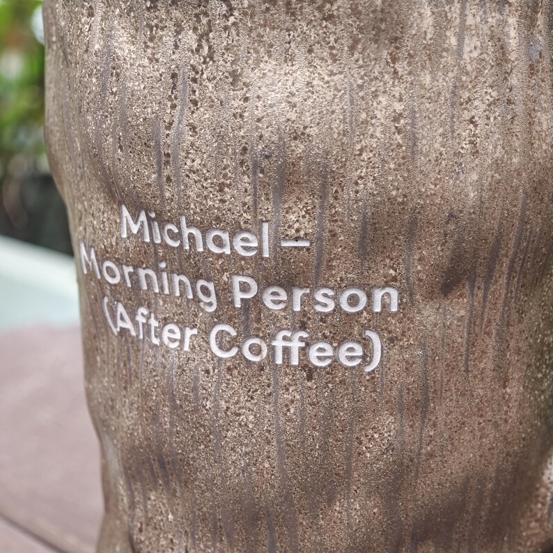 Puede incluir: Una taza cil&iacute;ndrica de color marr&oacute;n con una textura rugosa similar a la corteza de un &aacute;rbol. La superficie presenta un texto grabado en blanco que dice: Michael - Morning Person (After Coffee). El dise&ntilde;o imita un tronco con surcos verticales.