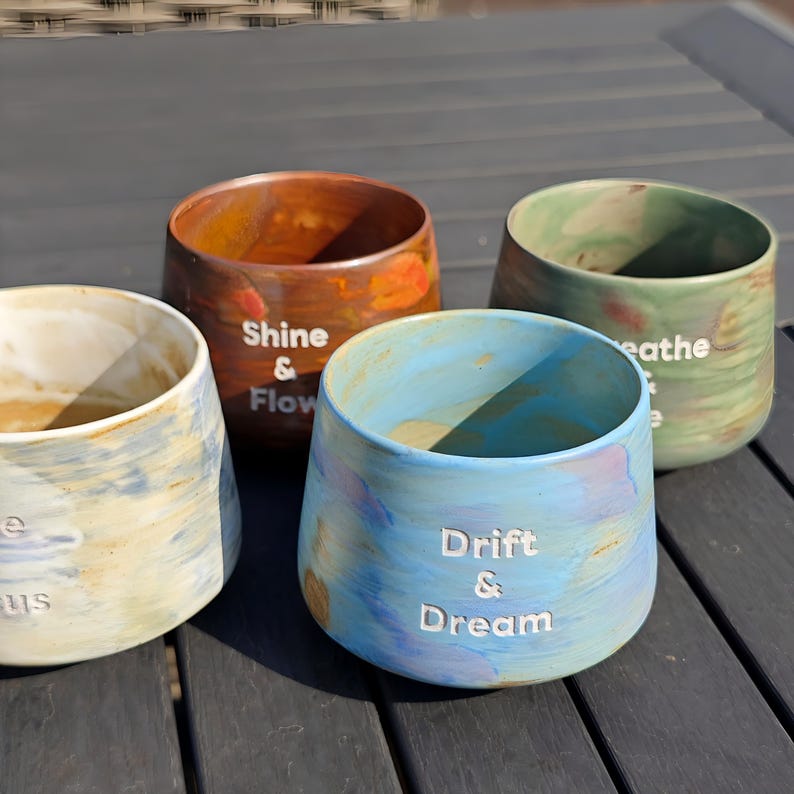 May include: Four handcrafted ceramic pots in shades of blue, brown, and green. Each pot has a unique design and text: "Shine & Flow", "Breathe", "Drift & Dream", and "Focus". The pots are on a dark wooden surface.