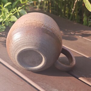Może przedstawiać: Okrągły ceramiczny kubek o teksturowanym, nakrapianym wykończeniu w odcieniach brązu i beżu. Kubek posiada zakrzywione ucho z boku oraz gładką, okrągłą podstawę. Powierzchnia ma nakrapiany wygląd z r&oacute;żnymi odcieniami ziemistego brązu na całości.