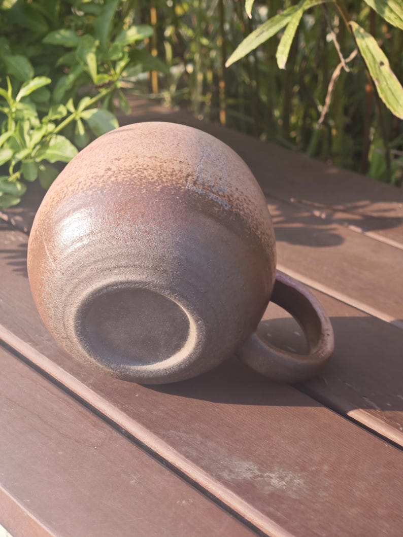 Może przedstawiać: Okrągły ceramiczny kubek o teksturowanym, nakrapianym wykończeniu w odcieniach brązu i beżu. Kubek posiada zakrzywione ucho z boku oraz gładką, okrągłą podstawę. Powierzchnia ma nakrapiany wygląd z r&oacute;żnymi odcieniami ziemistego brązu na całości.