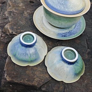 May include: A ceramic tea set with a blue, green, and white glaze. The set includes a lidded bowl, a saucer, and two small, flower-shaped cups. The tea set is placed on a dark, textured surface.