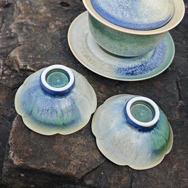 May include: A ceramic tea set with a blue, green, and white glaze. The set includes a lidded bowl, a saucer, and two small, flower-shaped cups. The tea set is placed on a dark, textured surface.