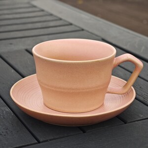 May include: A peach-colored ceramic teacup and saucer set. The cup has a square handle and a wide rim. The saucer has a ridged design. The set is made of a matte ceramic material.