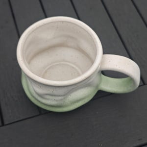 Op de afbeelding: Een handgemaakte keramische mok met een witte binnenkant en een gradi&euml;nt buitenkant die overgaat van wit naar zachtgroen. De mok heeft een afgeronde handgreep en een gespikkelde textuur, perfect voor koffie of thee.