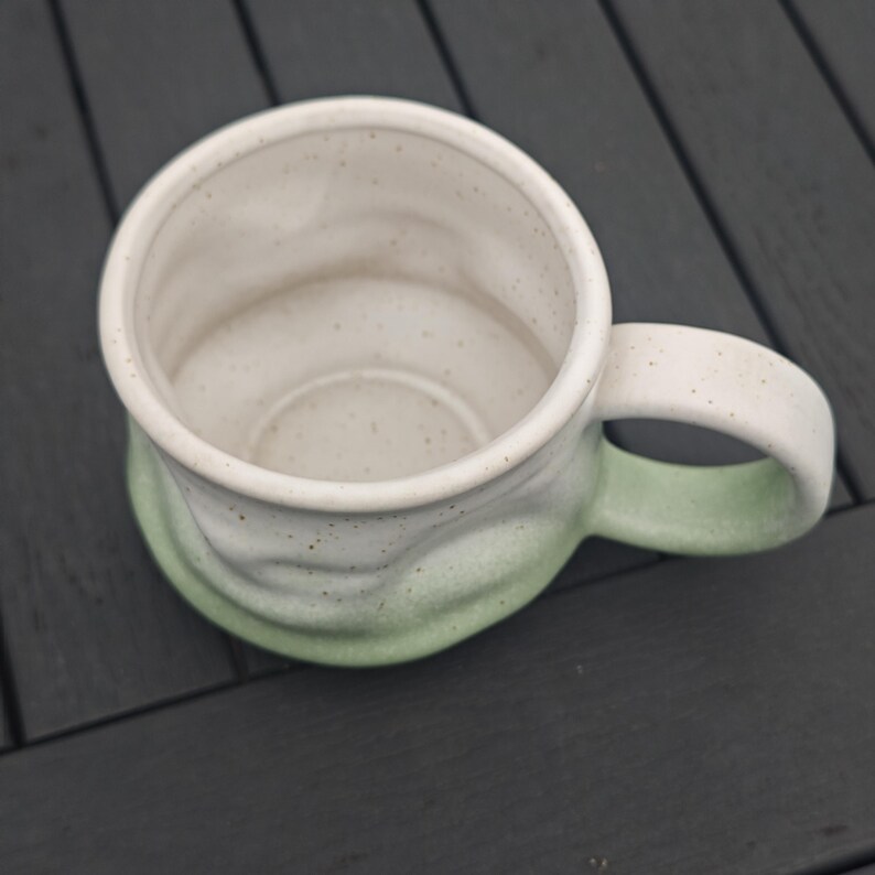 Op de afbeelding: Een handgemaakte keramische mok met een witte binnenkant en een gradi&euml;nt buitenkant die overgaat van wit naar zachtgroen. De mok heeft een afgeronde handgreep en een gespikkelde textuur, perfect voor koffie of thee.