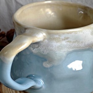 May include: A handmade ceramic mug with a unique design. The mug features a light blue base that transitions to a creamy beige rim. The handle is also beige. The mug's surface has a textured, glazed appearance, adding to its artisanal charm.