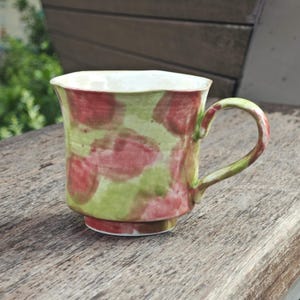 May include: A ceramic mug with a green and red spotted pattern. The mug has a white interior and a rounded handle. The mug is sitting on a wooden surface. The mug is approximately 4 inches tall.